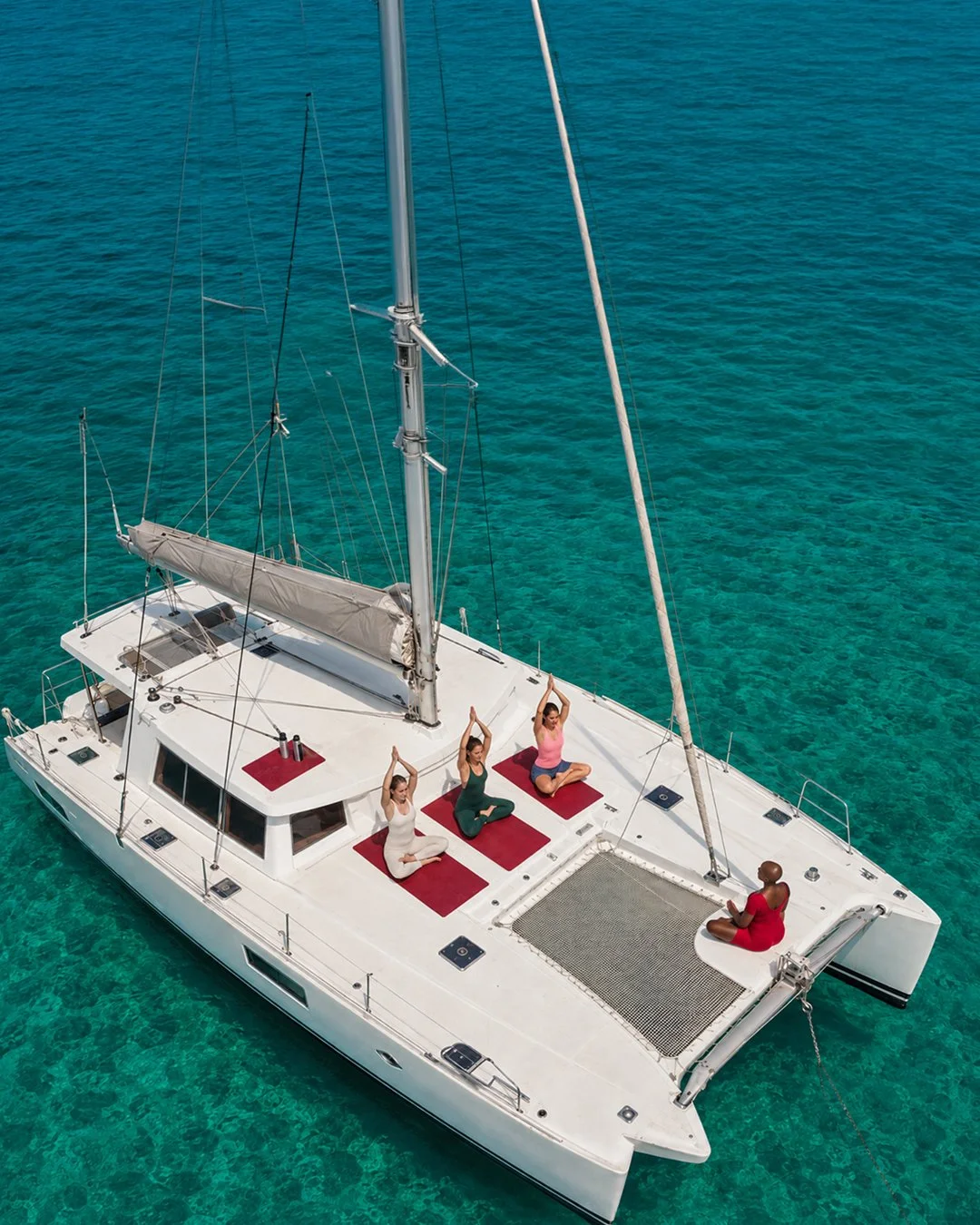 Four women practicing yoga on the deck of a white sailboat with red mats, floating on clear blue water.
