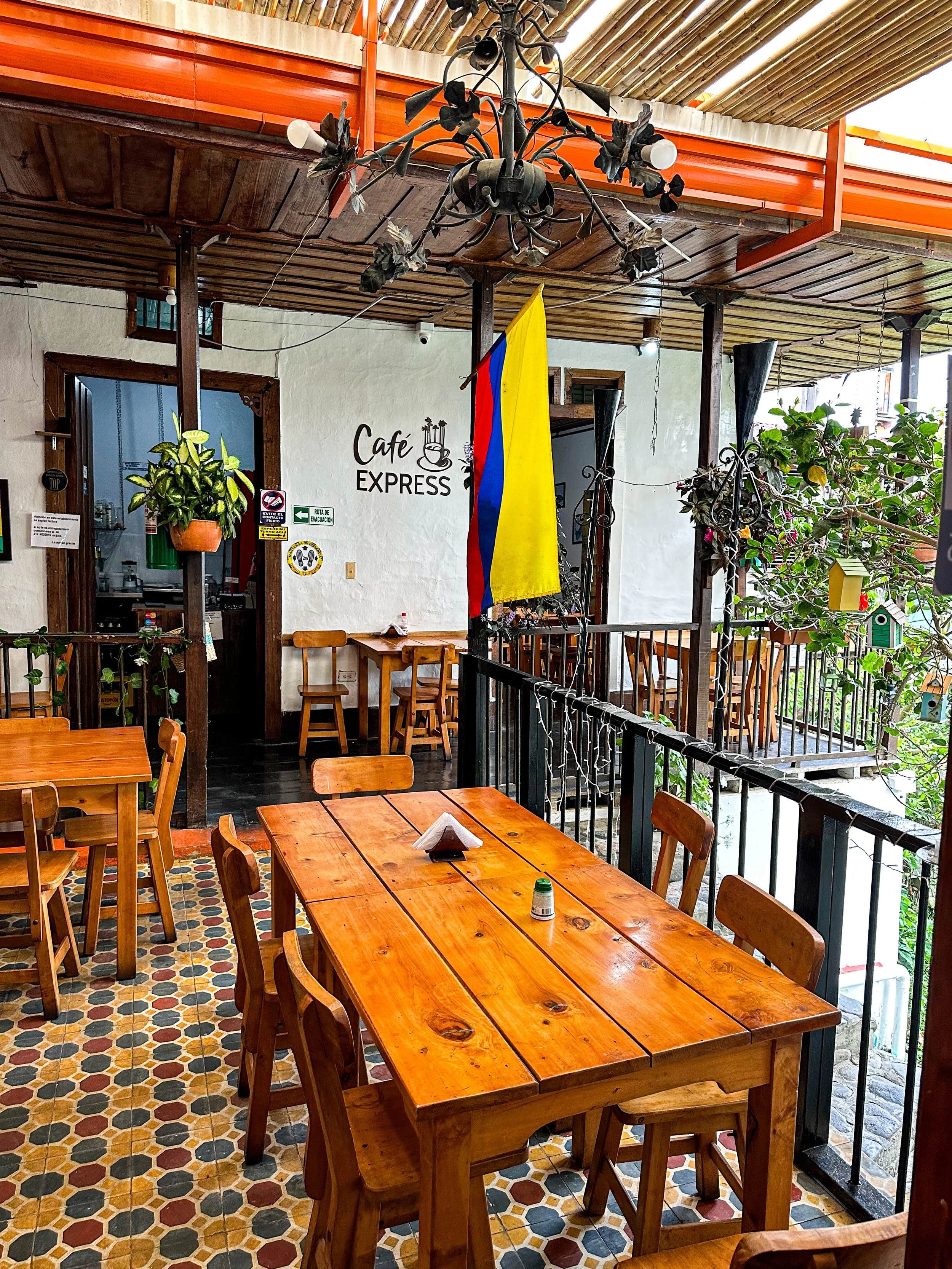 Outdoor seating area of a café with wooden tables and chairs, a Colombian flag hanging, and decorative plants, with a sign reading "Café Express" on the wall.