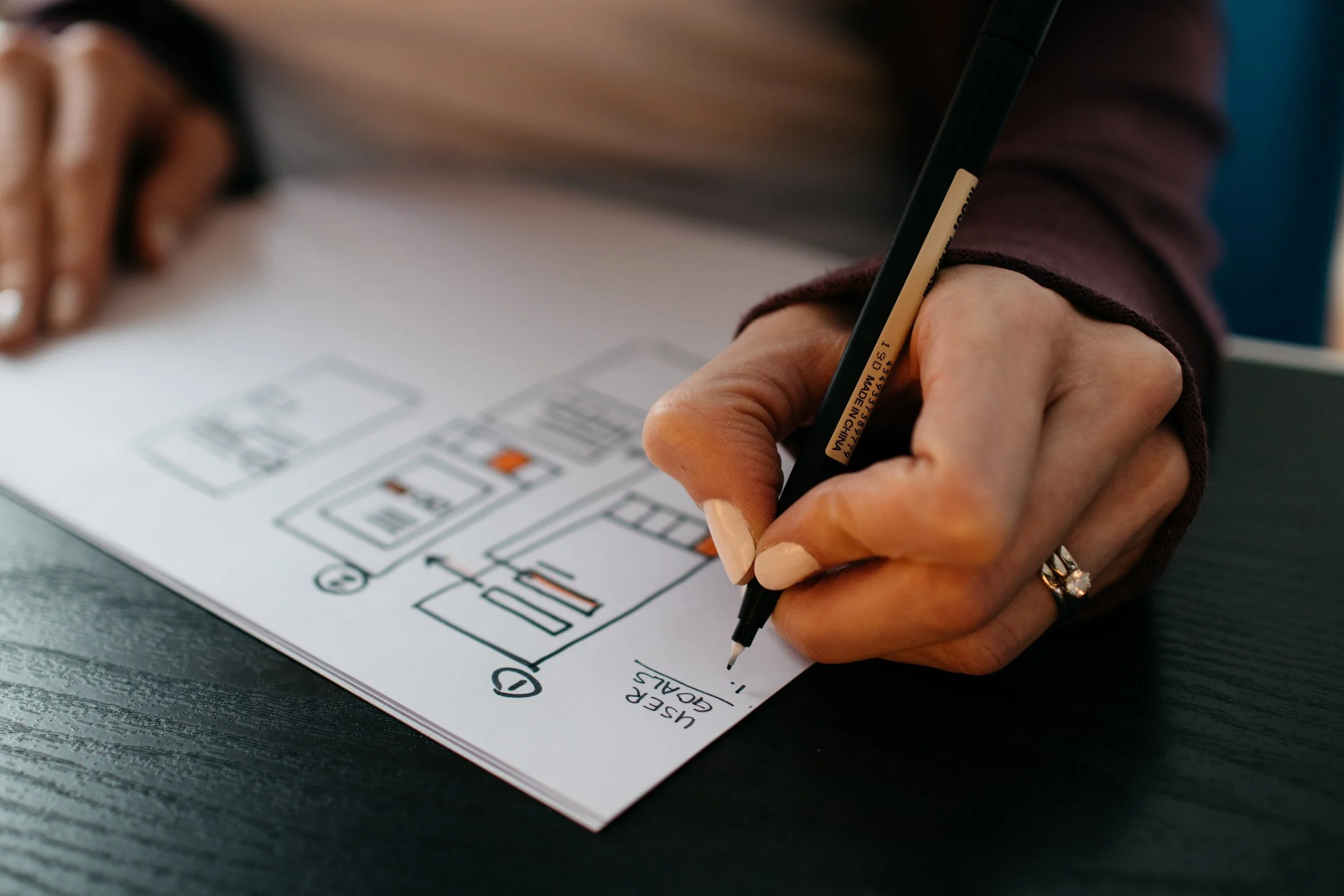 A person is writing on a sheet of paper with diagrams and labeled sections, using a black pen, on a dark tabletop.