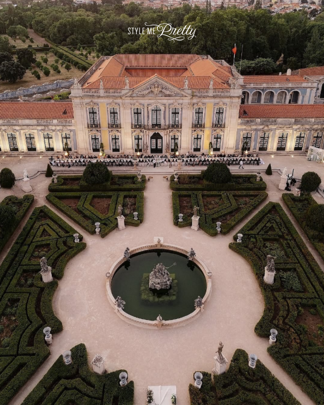 An aerial view of a grand, historic building with a red tiled roof, surrounded by manicured gardens, statues, and a fountain in the center, with a cityscape and trees in the background.