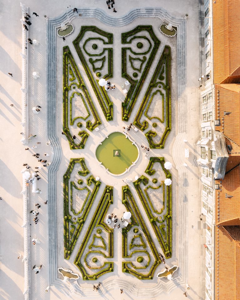 Overhead view of a symmetrical garden with hedges, pathways, and a central fountain, surrounded by buildings and people walking nearby.