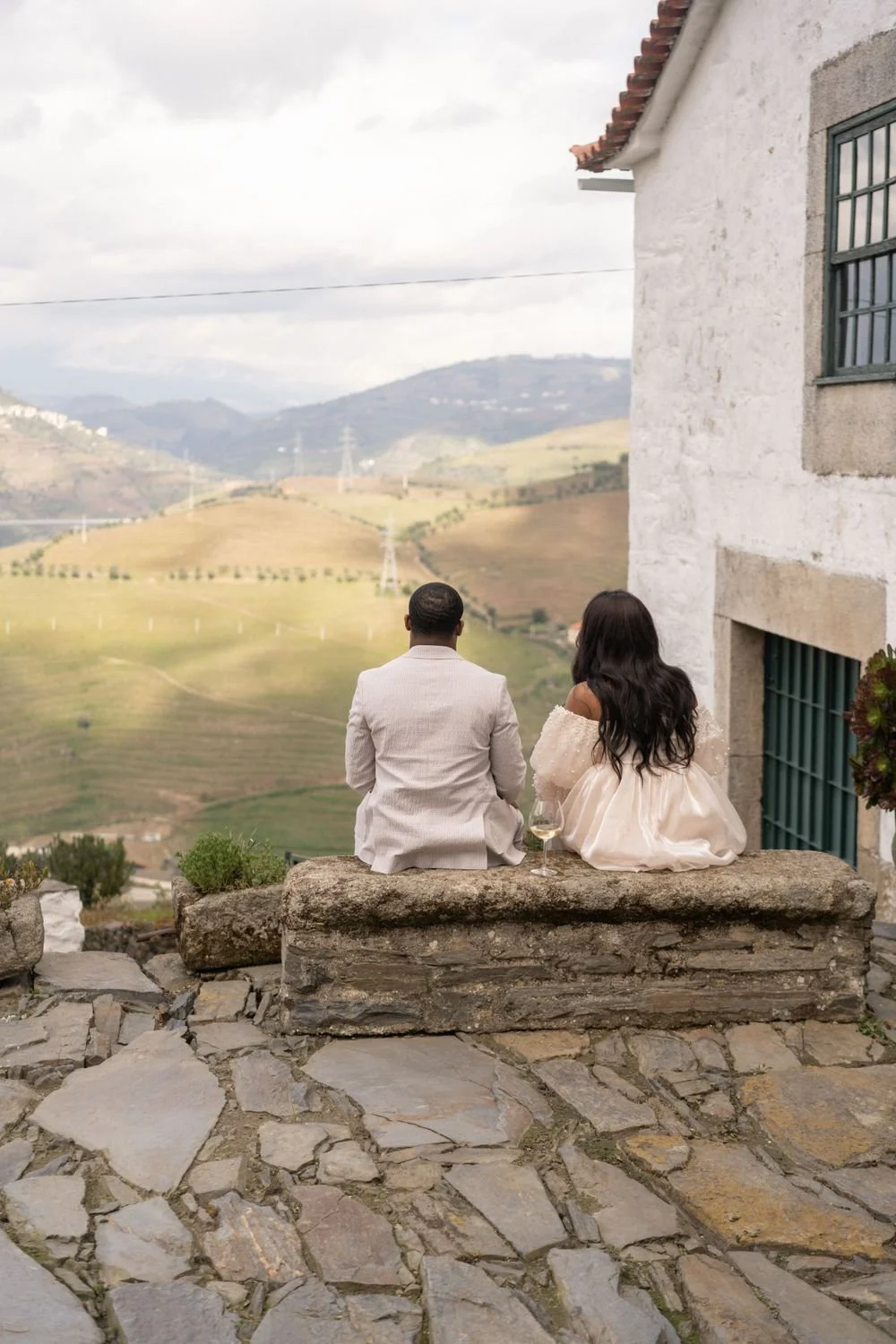 A man and woman dressed in formal attire, sitting on a stone ledge overlooking rolling hills and fields, with a glass of white wine between them, in a rustic outdoor setting.
