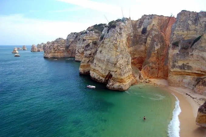 Cliffside view of a beach with turquoise water, sandy shoreline, and towering sandstone cliffs with layered formations.