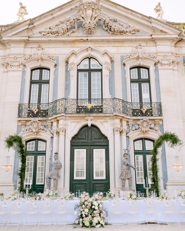 The facade of an ornate, historic building with decorative stucco details, tall windows, and a balcony with intricate wrought iron railing. There is a long table set for a formal event in front, decorated with flowers and greenery, flanked by statues of knights or soldiers.