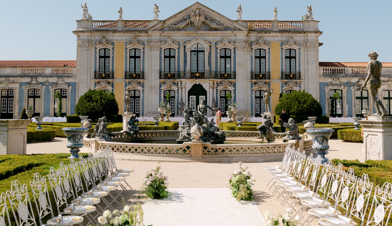 Luxurious historic palace with ornate facade and classical statues in front, surrounded by manicured gardens and seating area, possibly prepared for an outdoor event.