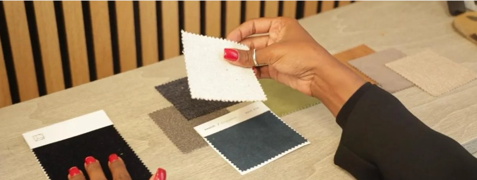 Person compares fabric swatches on a table, holding a white fabric piece in one hand.