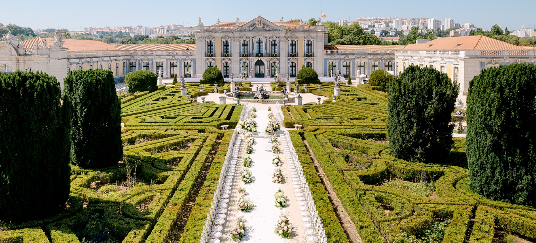 View of a grand palace with elaborate gardens featuring neatly trimmed hedges, fountains, sculptures, and a pathway with floral arrangements leading to the entrance of the palace.