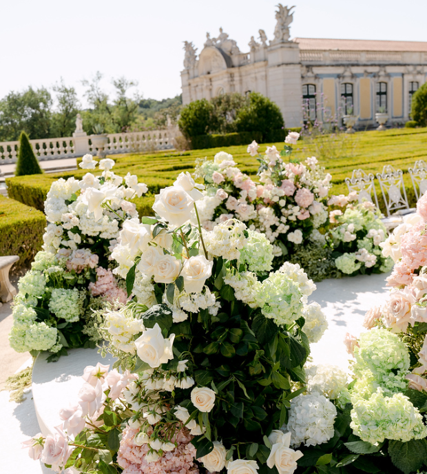 Elegant outdoor garden with white and pink flower arrangements, classical building in the background, on a sunny day.