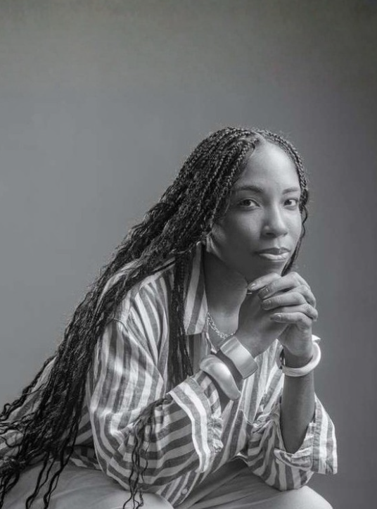 Black and white portrait of a woman with long braided hair, wearing a striped shirt, with her hands clasped near her face, looking at the camera with a slight smile.