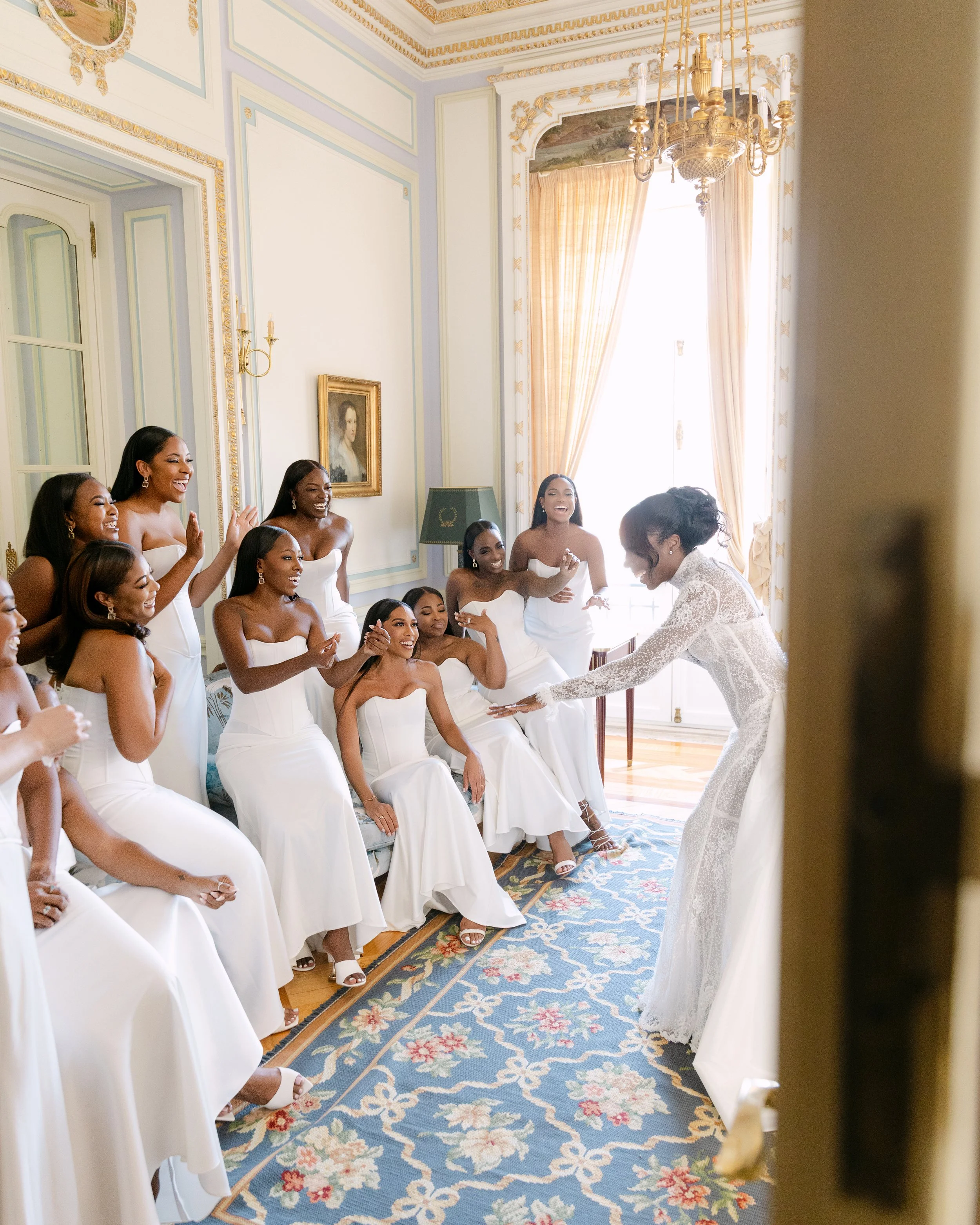 Bride in lace dress entertaining her bridesmaids in a elegant room with large window, floral rug, and gold accents.