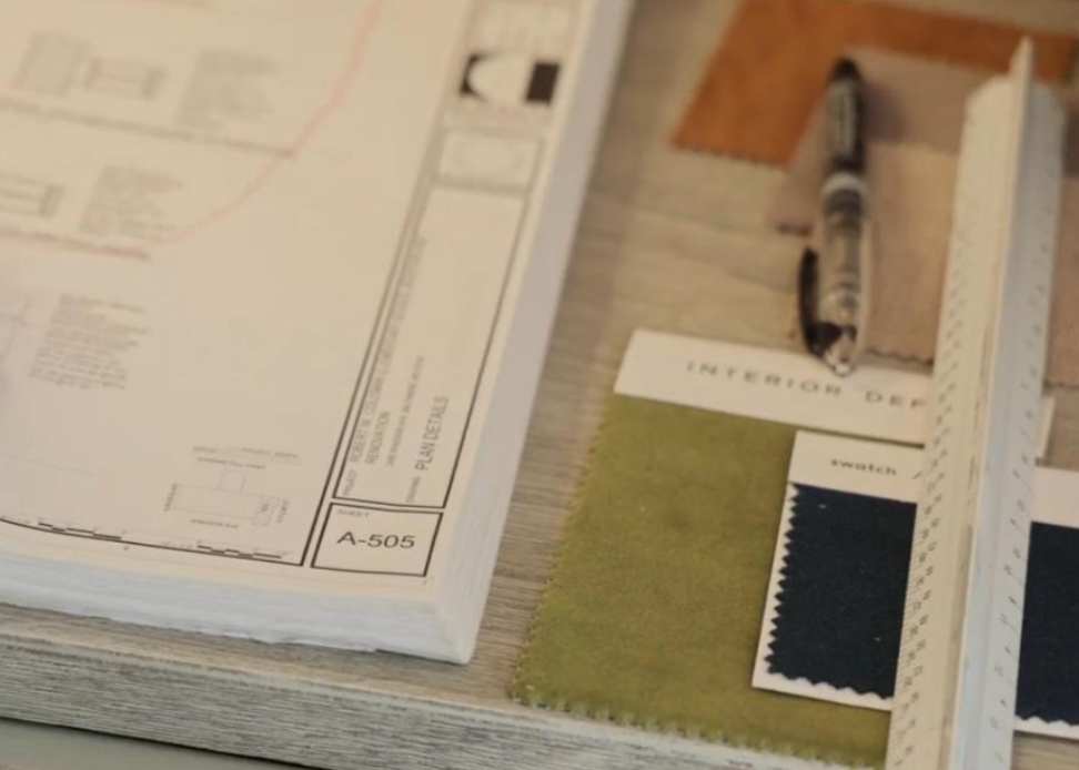 Architectural plans, a marker, a ruler, color swatches, and fabric samples on a wooden table.