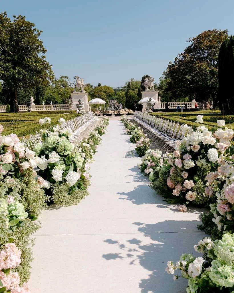 A wedding aisle decorated with white and pastel flowers, leading to sculptures and statues in an outdoor garden setting under a clear blue sky.