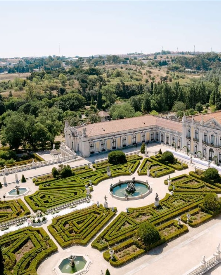 Aerial view of a formal garden with symmetrical hedges, fountains, and sculptures in front of a large historic building, surrounded by trees and a countryside landscape.