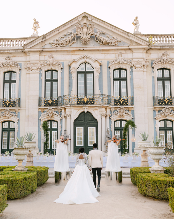 Bride and groom walking towards a grand, ornate mansion with statues and decorative architectural details, flanked by two tall, dressed-up figures on stilts in white gowns holding bouquets.