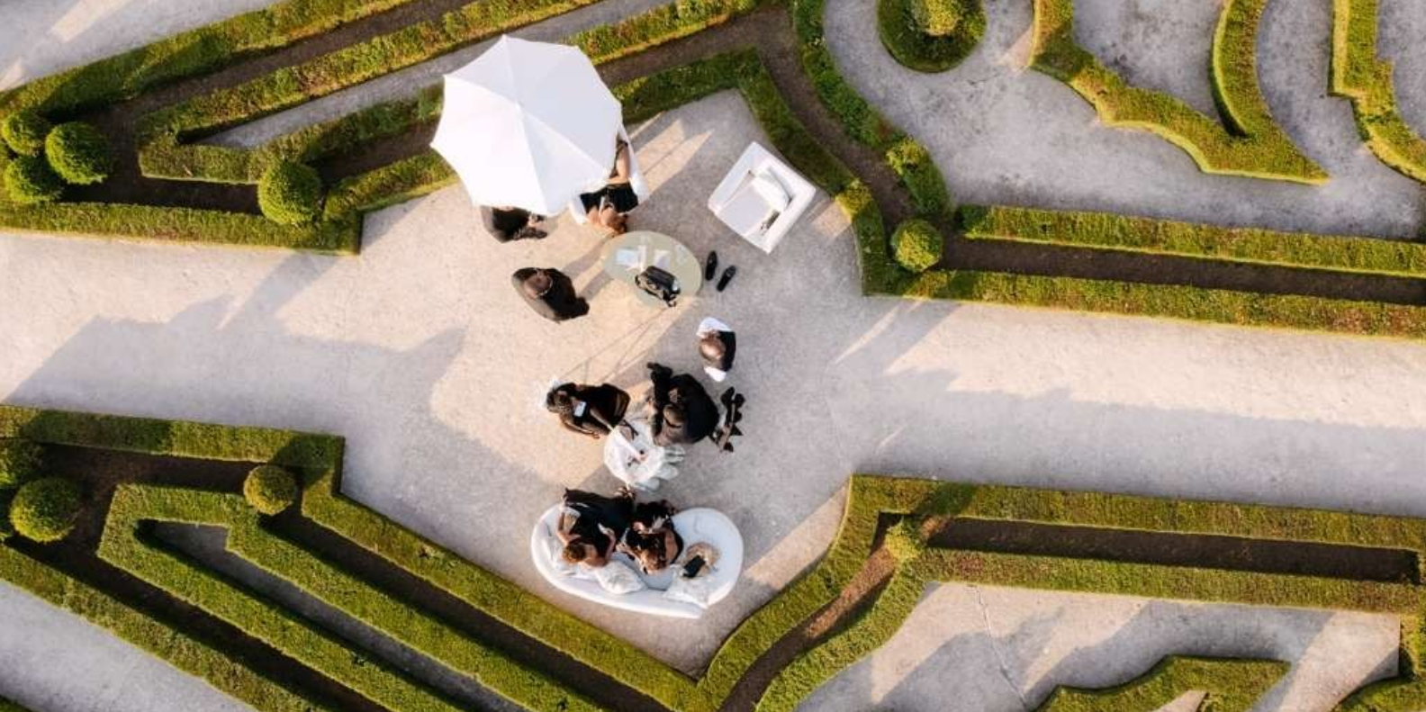 People gathered around a table with a white umbrella outdoors in a garden or park, seen from above.