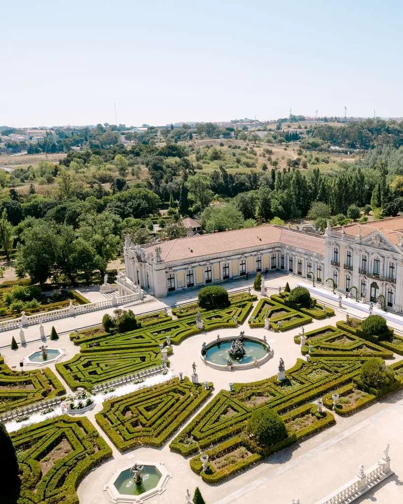 Aerial view of a large, ornate mansion with extensive, geometrically designed garden and fountains, surrounded by lush greenery and rolling hills.