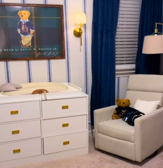 A teddy bear sitting on a white armchair next to a window with blue curtains, a dresser with a changing pad on top, and a framed poster on the wall in a cozy room.