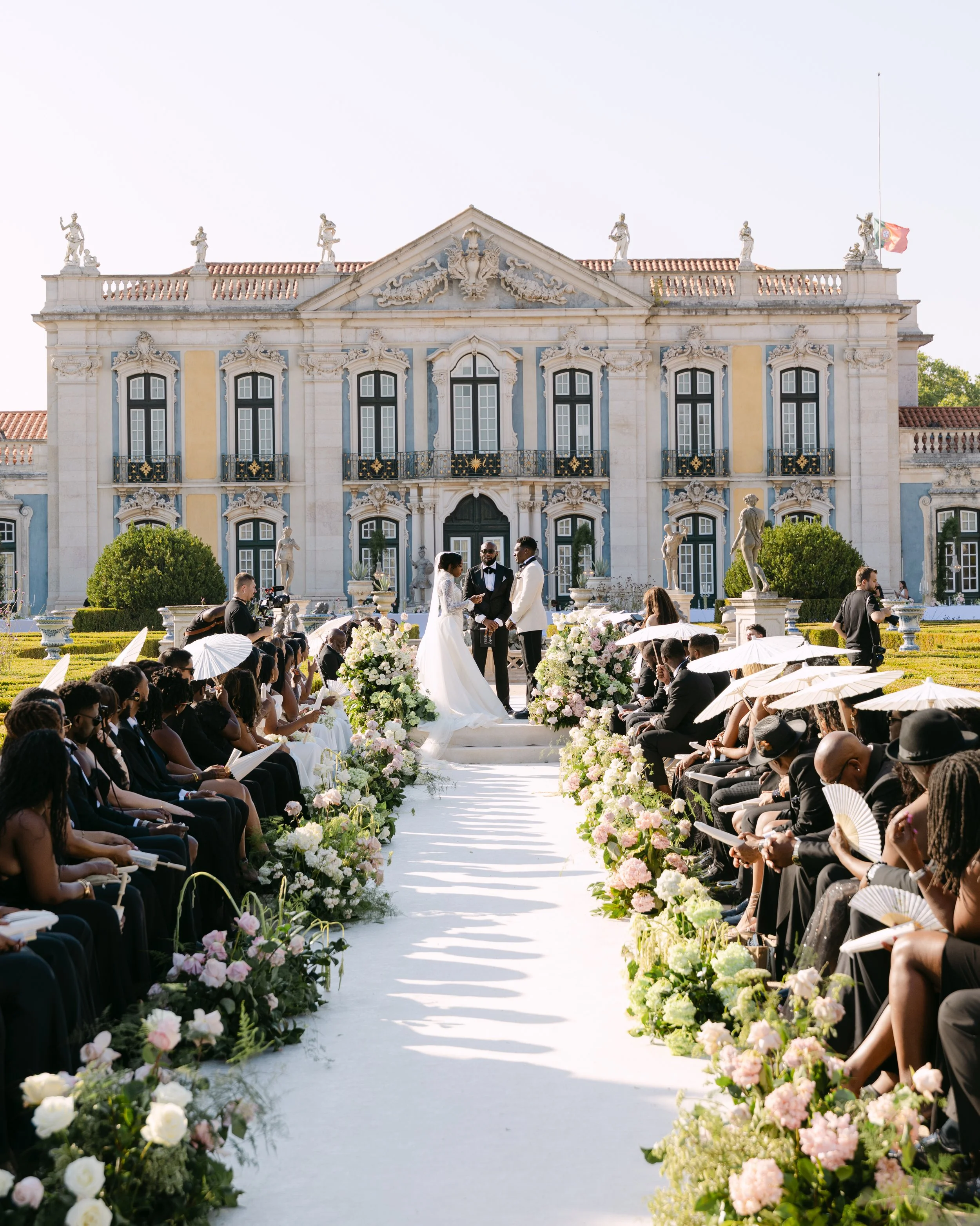 Featured in Vogue: Architect-Led Luxury. A wedding ceremony taking place outdoors in front of a large, ornate mansion. The bride and groom are standing at the altar with a minister, with guests seated on either side.