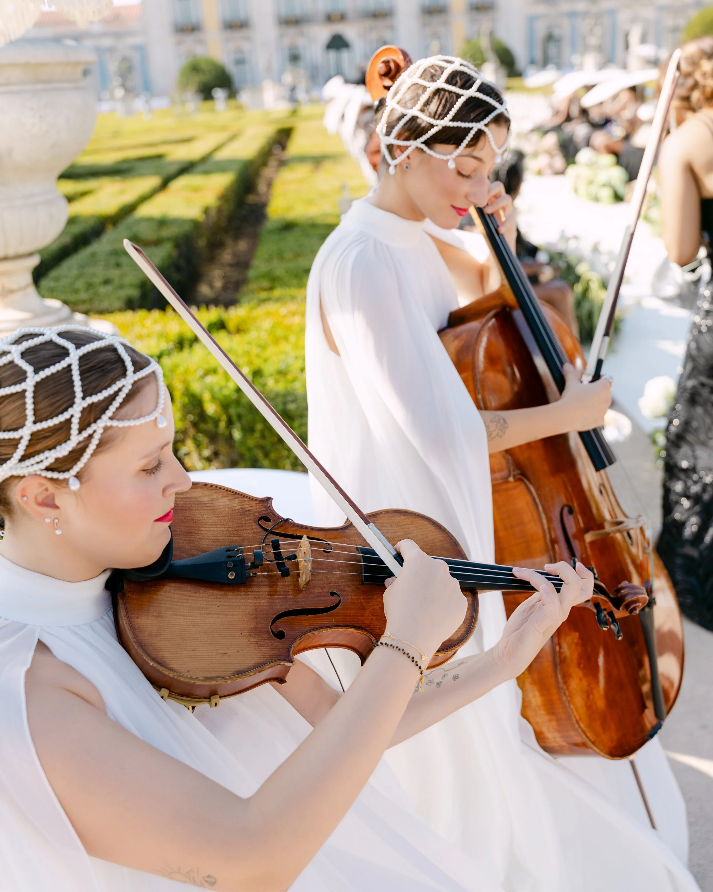 Two women in white dresses playing violins outdoors, wearing decorative headpieces, with a garden and other people in the background.