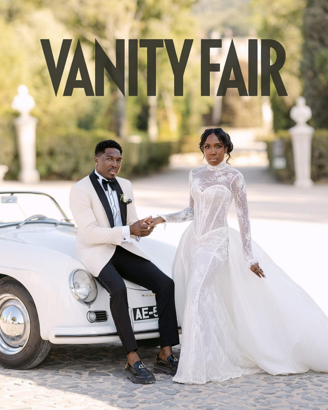 A bride and groom in wedding attire sitting beside a vintage white convertible, holding hands outdoors with trees and decorative pillars in the background, at a Vanity Fair event or photoshoot.