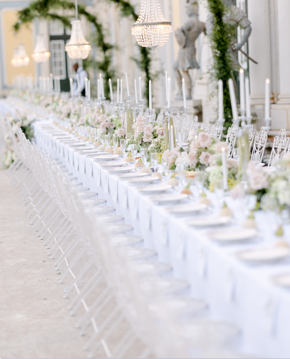 Long banquet table set for an event with white tablecloths, floral centerpieces, and tall candlesticks, decorated with chandeliers and elegant decor.