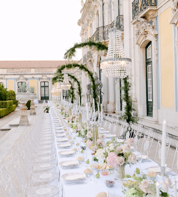 Elegant outdoor dining table set for a formal event with white tablecloth, floral centerpieces, and chandeliers, on a patio of a historic building with ornate architecture.