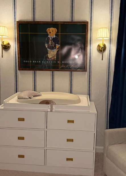 A white changing table with a baby bathtub on top, situated in a room with striped wallpaper, two wall-mounted lights, a framed poster of a Polo Bear dressed in clothing, and a partly visible armchair on the right.