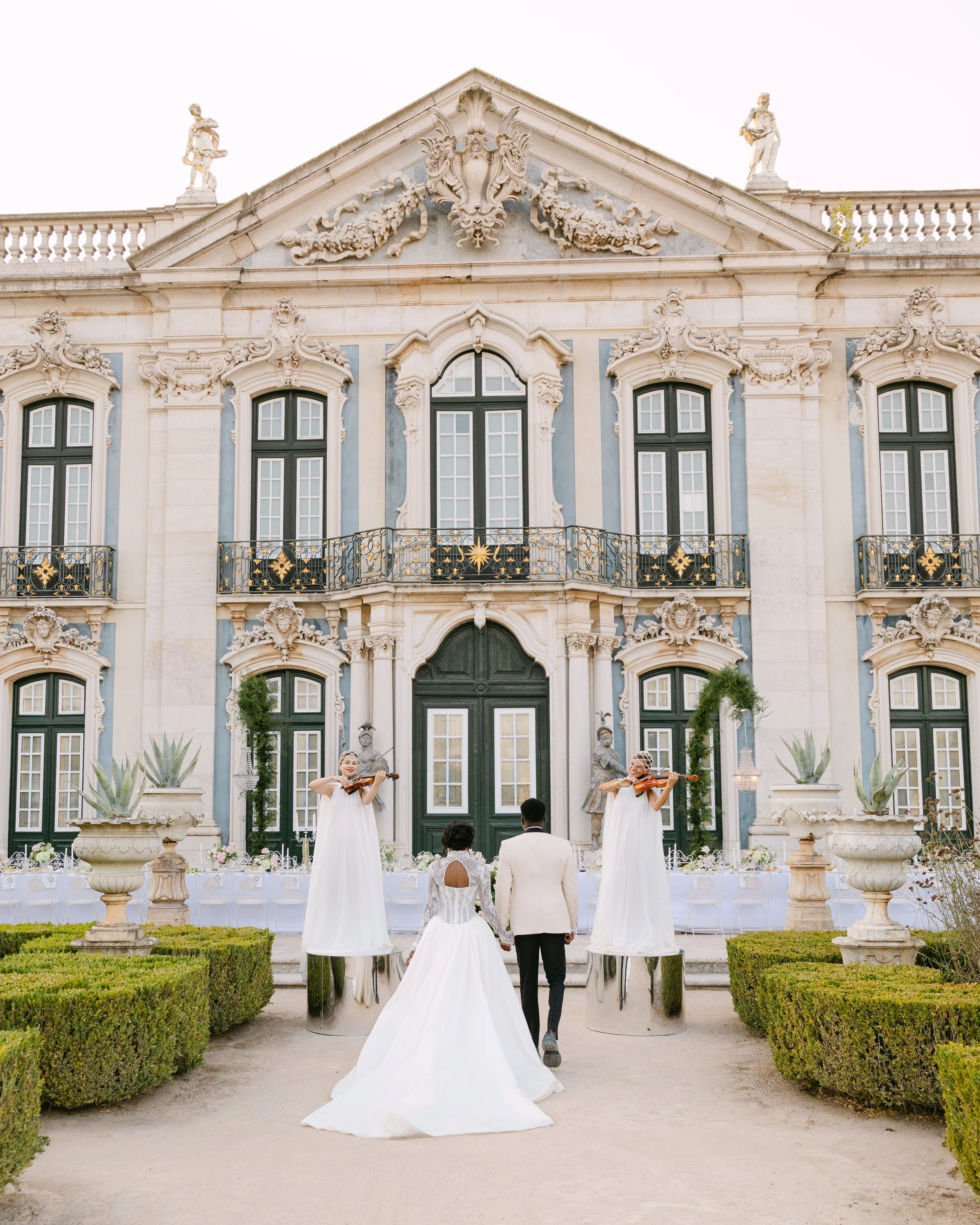 National Palace of Queluz Wedding - Bride and groom walk towards a grand, ornate mansion with two women playing violins on elevated platforms in front of the entrance.