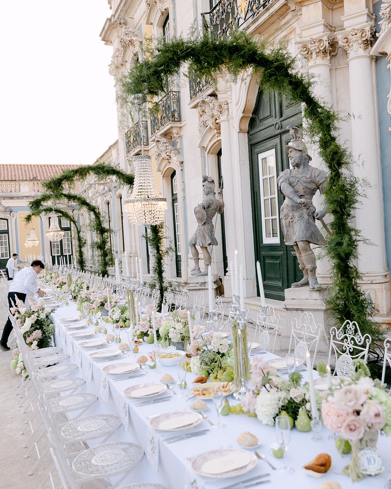 Elegant outdoor wedding setup with a long table decorated with white linens, floral arrangements, candles, and greenery. Surrounding the table are white ornate chairs, and a building with classical architecture and statues serves as the backdrop. Ove