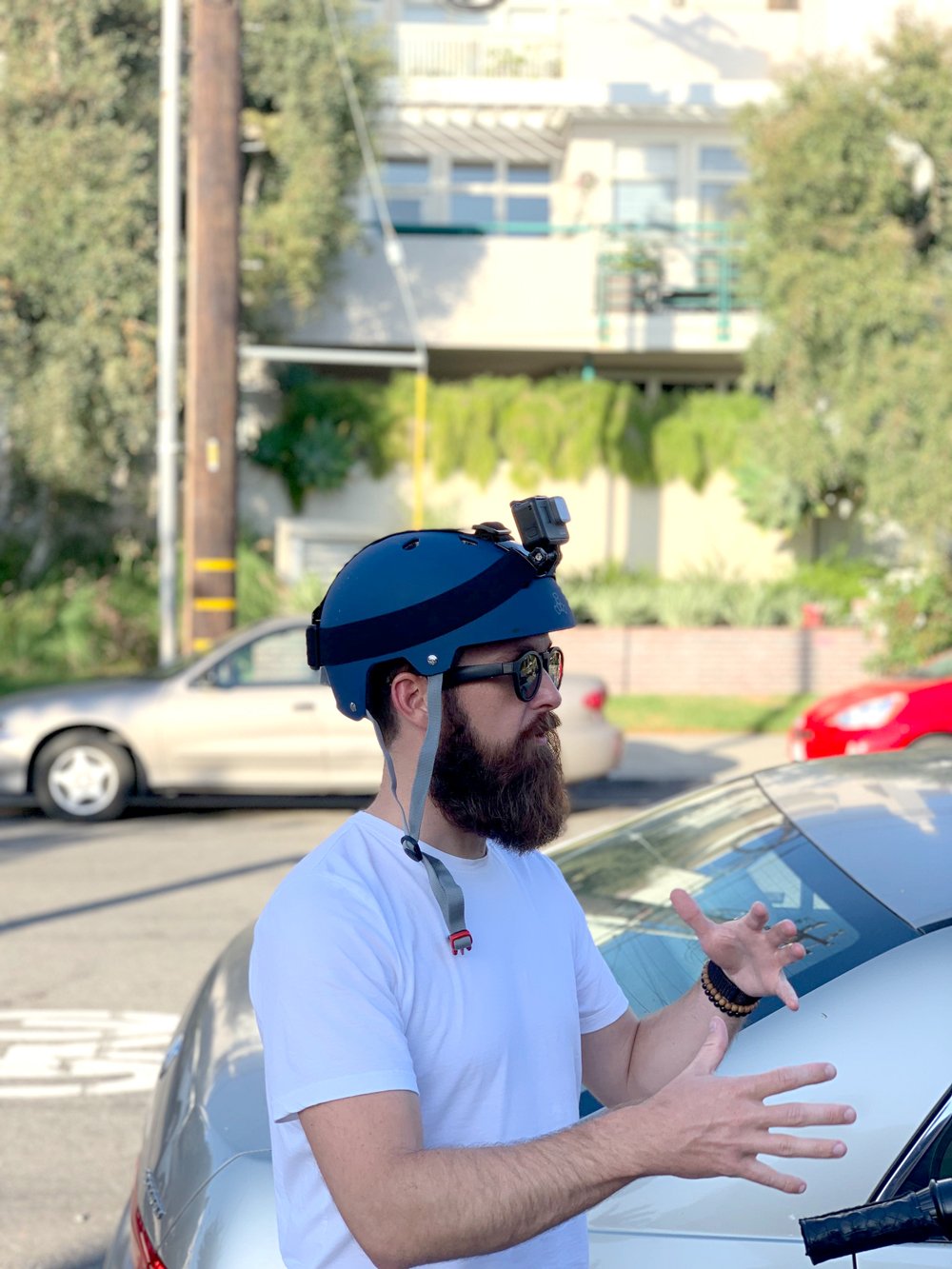 A man wearing a helmet with a mounted camera gestures while speaking outdoors