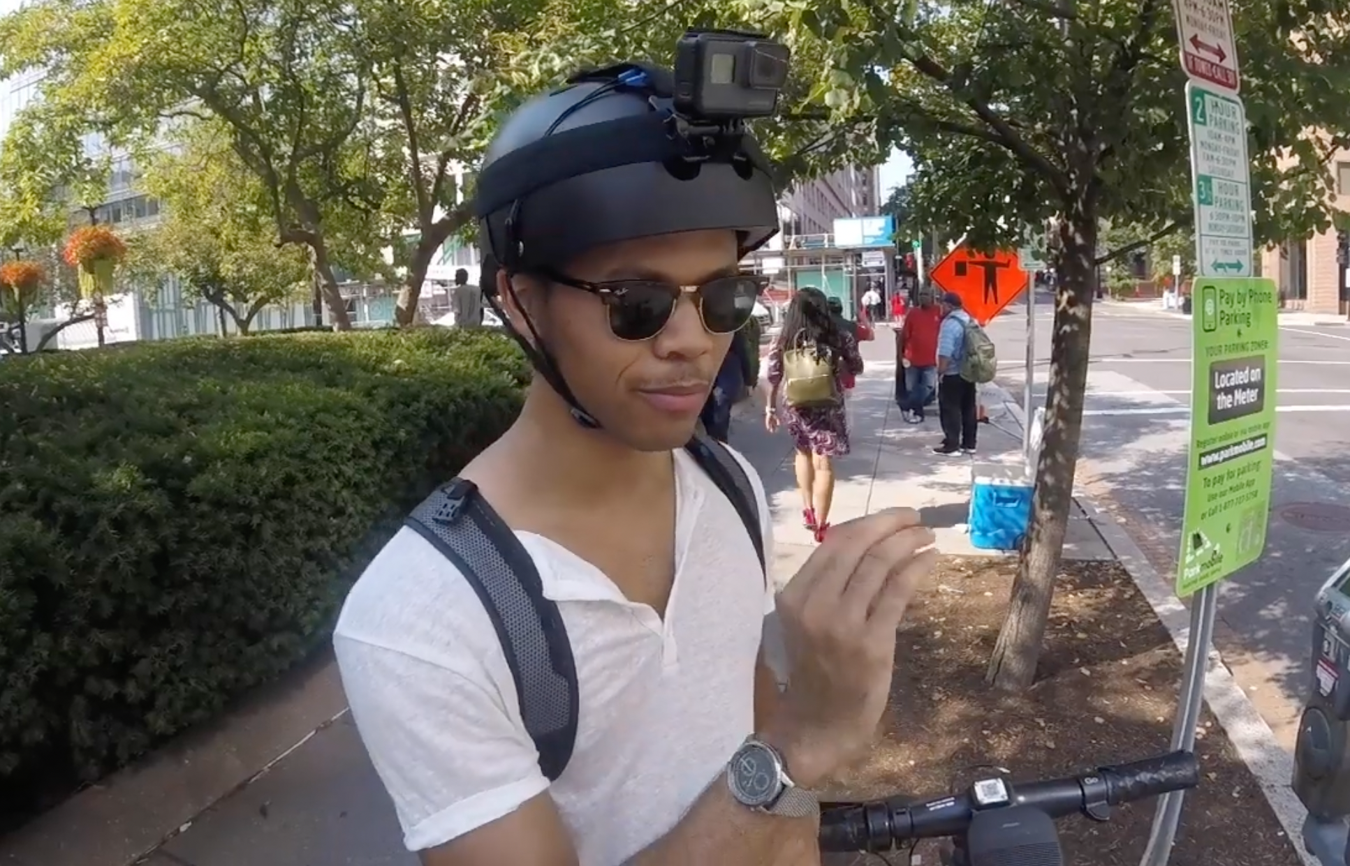 A person wearing a helmet with a mounted camera, holding a scooter handlebar at a street corner