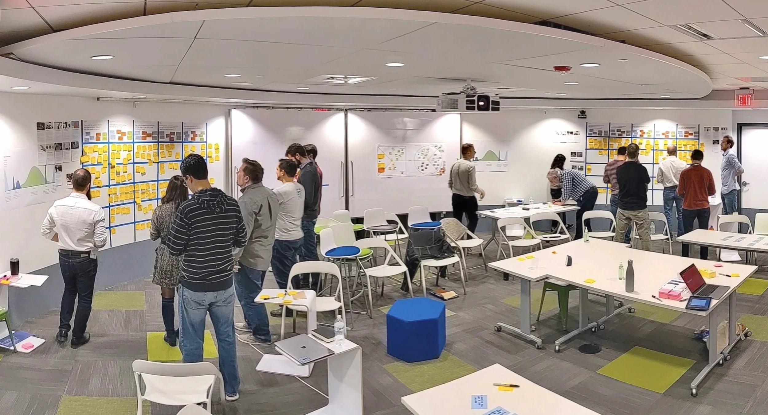 A panoramic shot of several people in a modern workshop space, gathered around whiteboards filled with sticky notes and diagrams