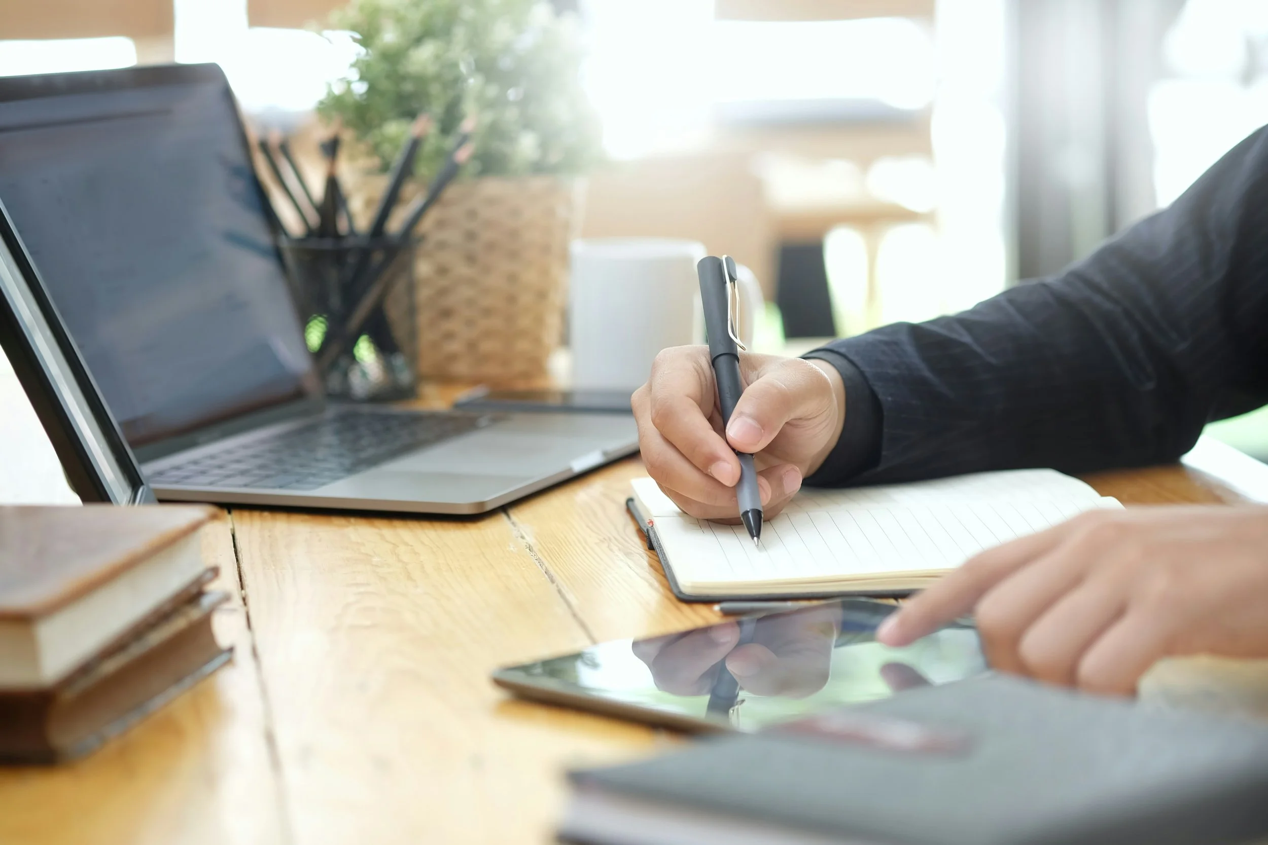 A person writing in a notebook with a pen while also using their other hand to interact with a tablet