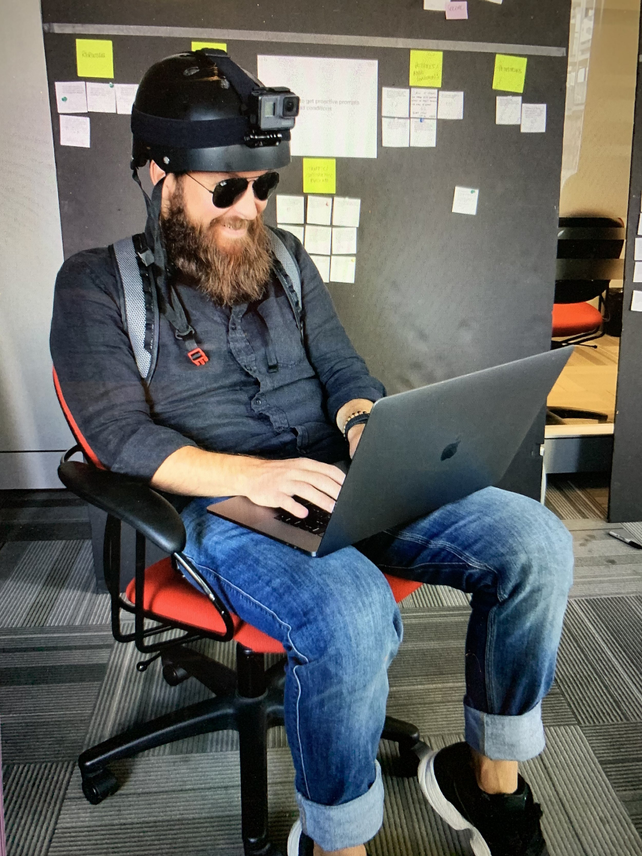 A man sitting at a desk wearing a helmet with a camera mounted on it, working on a laptop