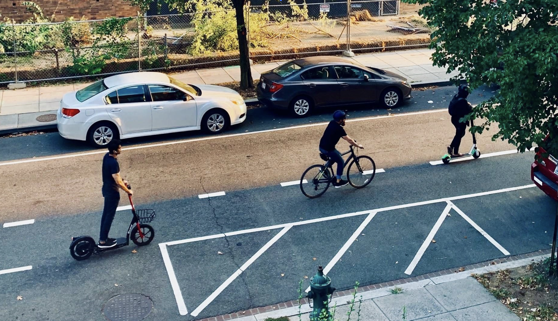 A street with a cyclist, a person on an electric scooter, and another person on a motorized kick scooter, with parked cars in the background