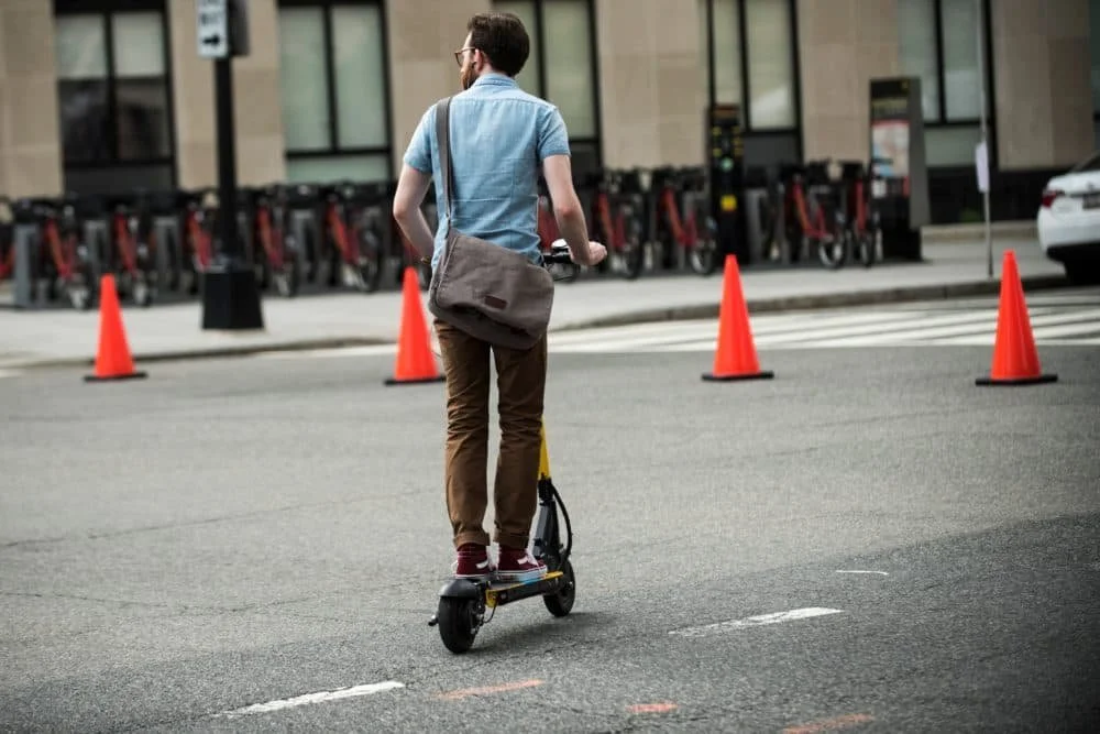 A man on an electric scooter with a bag, riding near orange cones and parked bike