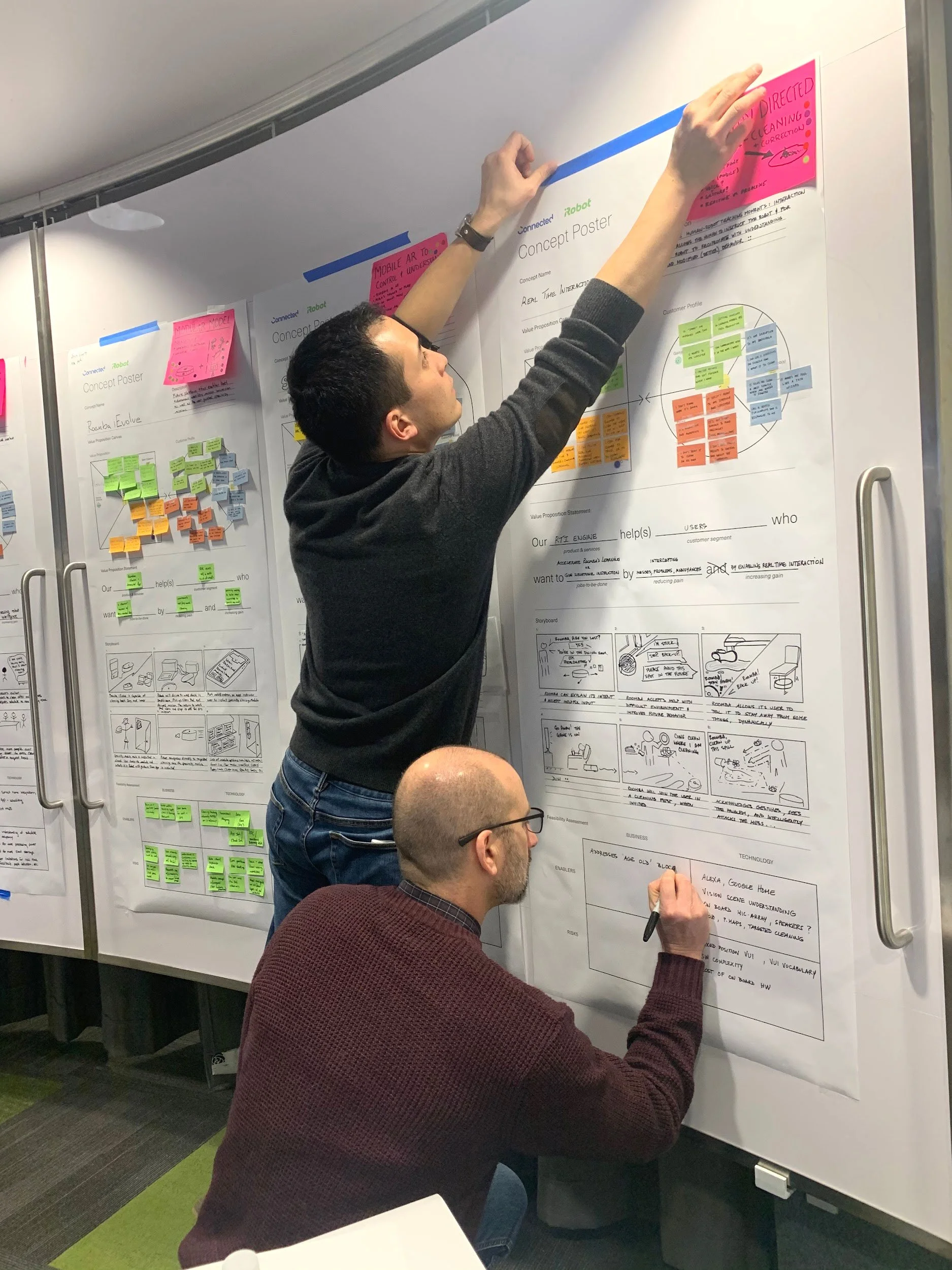 A person kneeling and writing on a storyboard sheet attached to a whiteboard while another person adds content above
