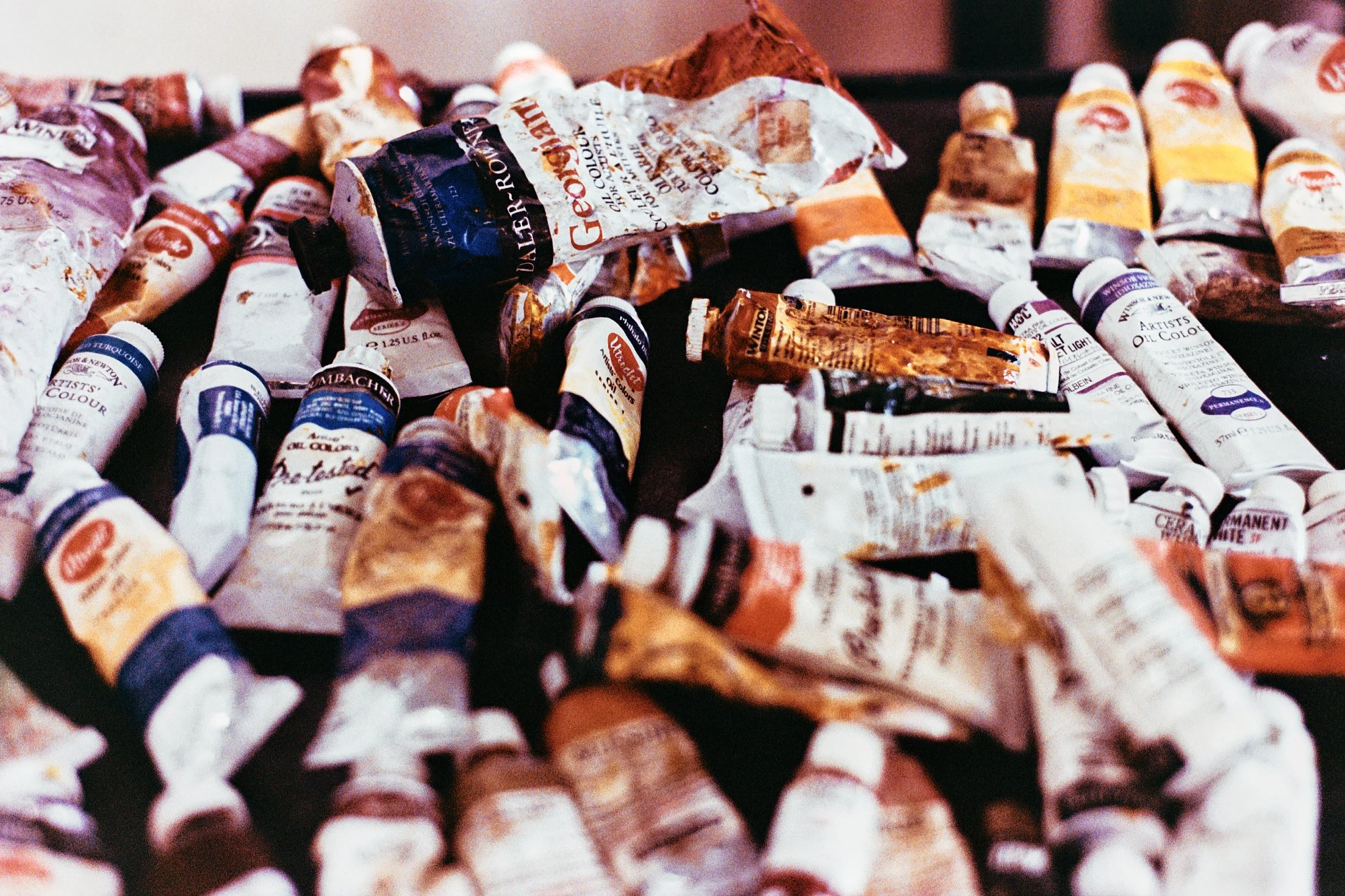 A pile of used oil paint tubes, some empty and crumpled, with paint stains, labels, and caps, scattered across a dark surface.