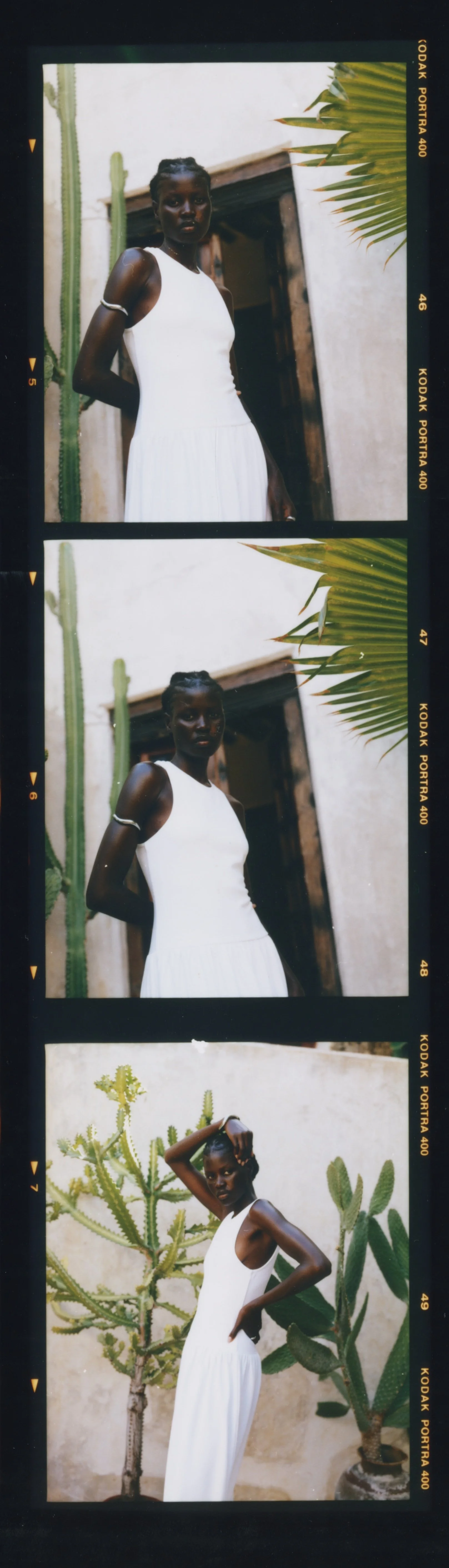 A young woman with dark skin and braided hair, wearing a white sleeveless dress, standing outdoors near cactus plants and a white wall, with a wooden door in the background.