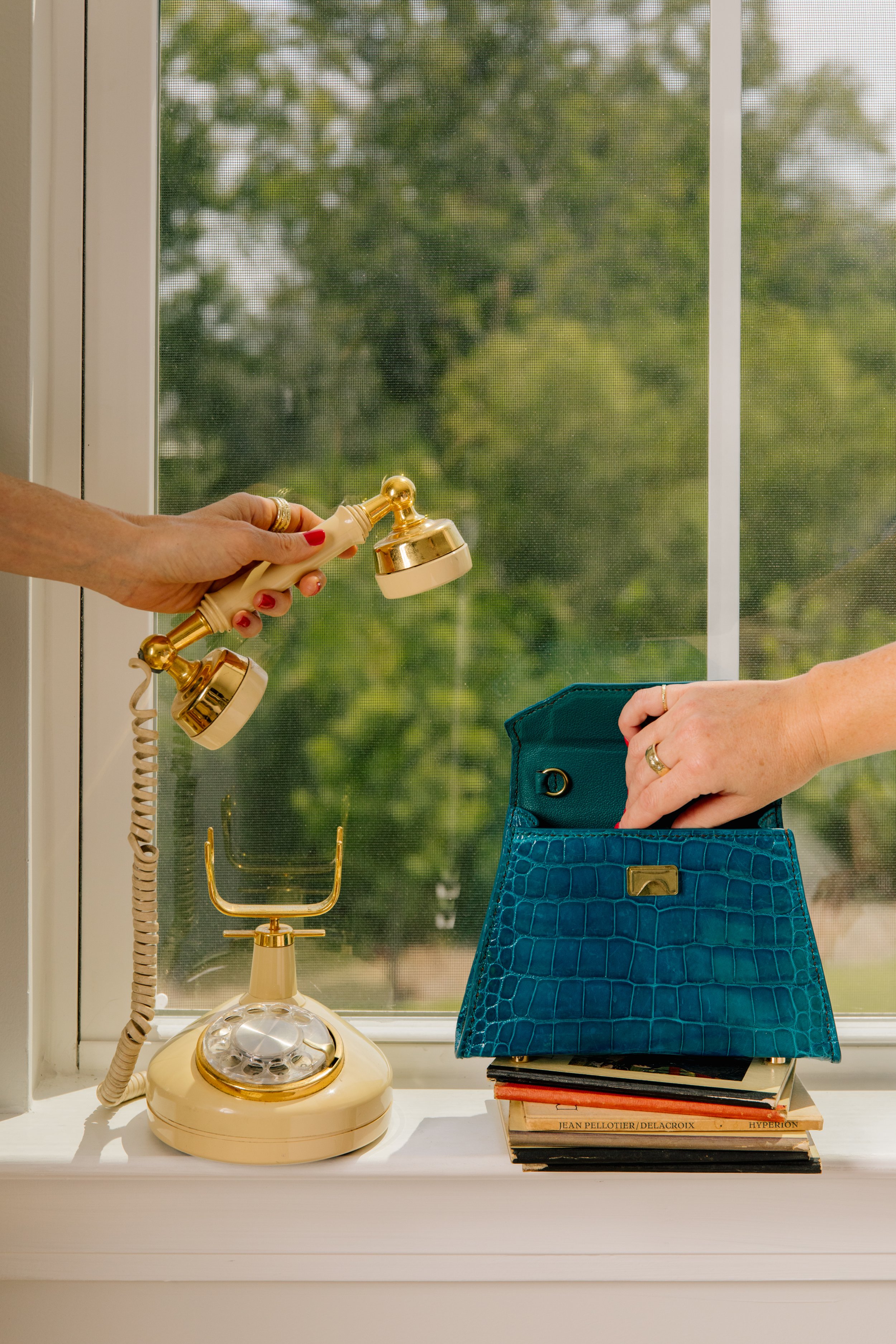 A vintage cream-colored rotary telephone on a windowsill with a golden handle. A person is reaching into a teal leather purse placed on a stack of magazines, with book titles visible.