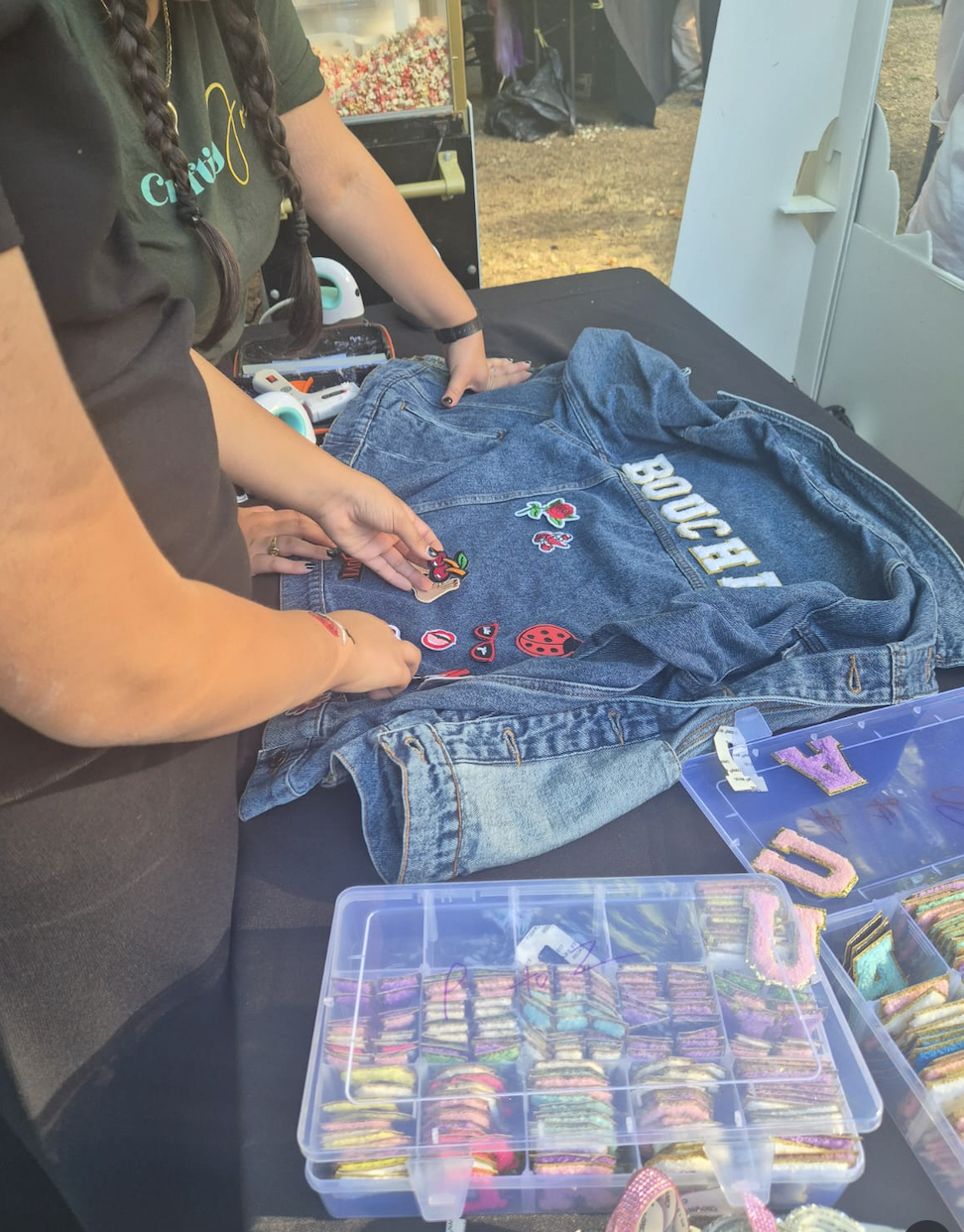 People decorating a denim jacket with patches at a craft fair or outdoor event. There are containers of colorful patches and letters on the table.