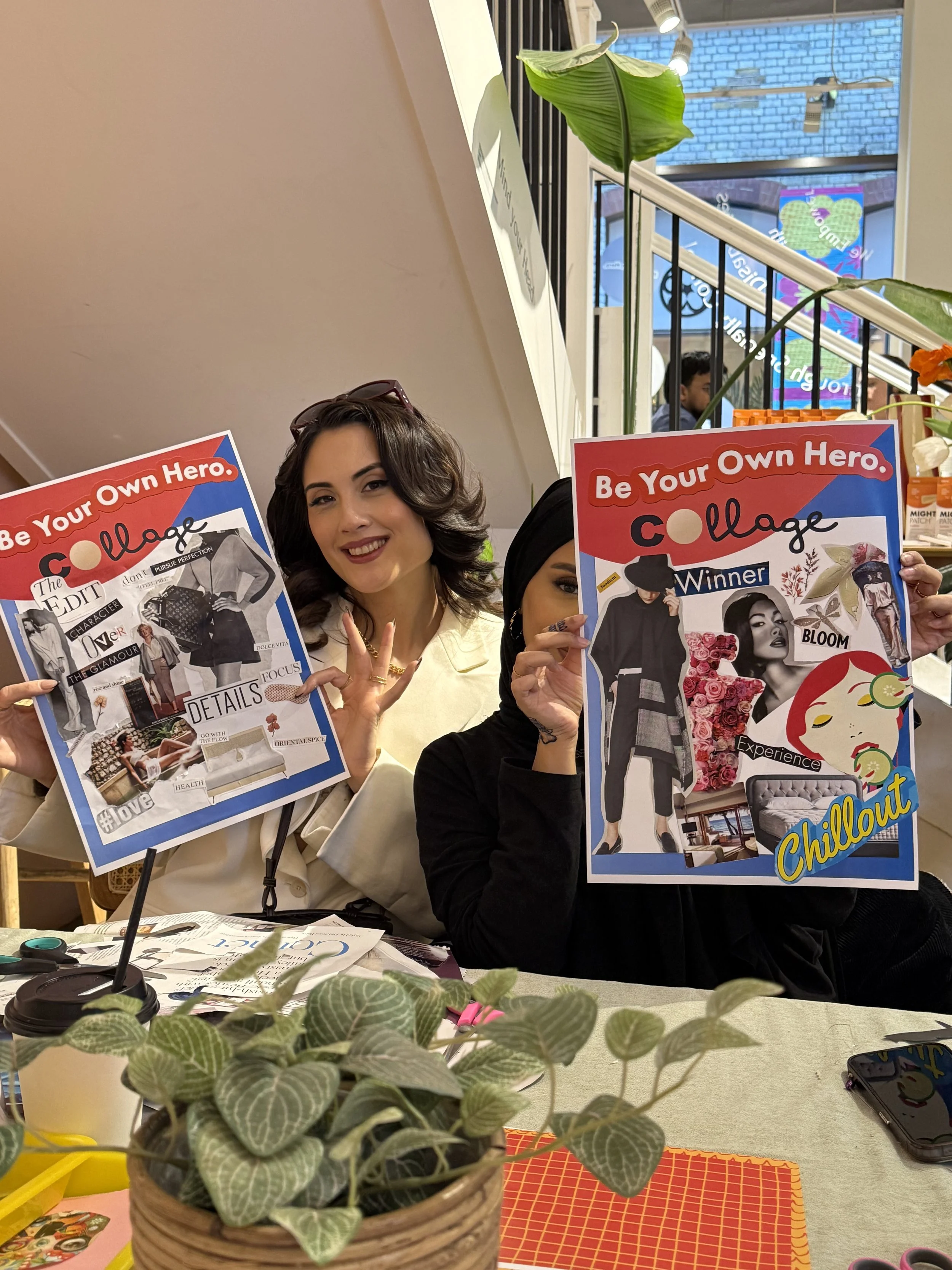 Two women sitting at a table, holding up colorful posters promoting a collage activity. One woman with dark hair and a beige blazer smiles at the camera. The other woman, wearing a black hijab and dark clothing, holds her poster partially covering he