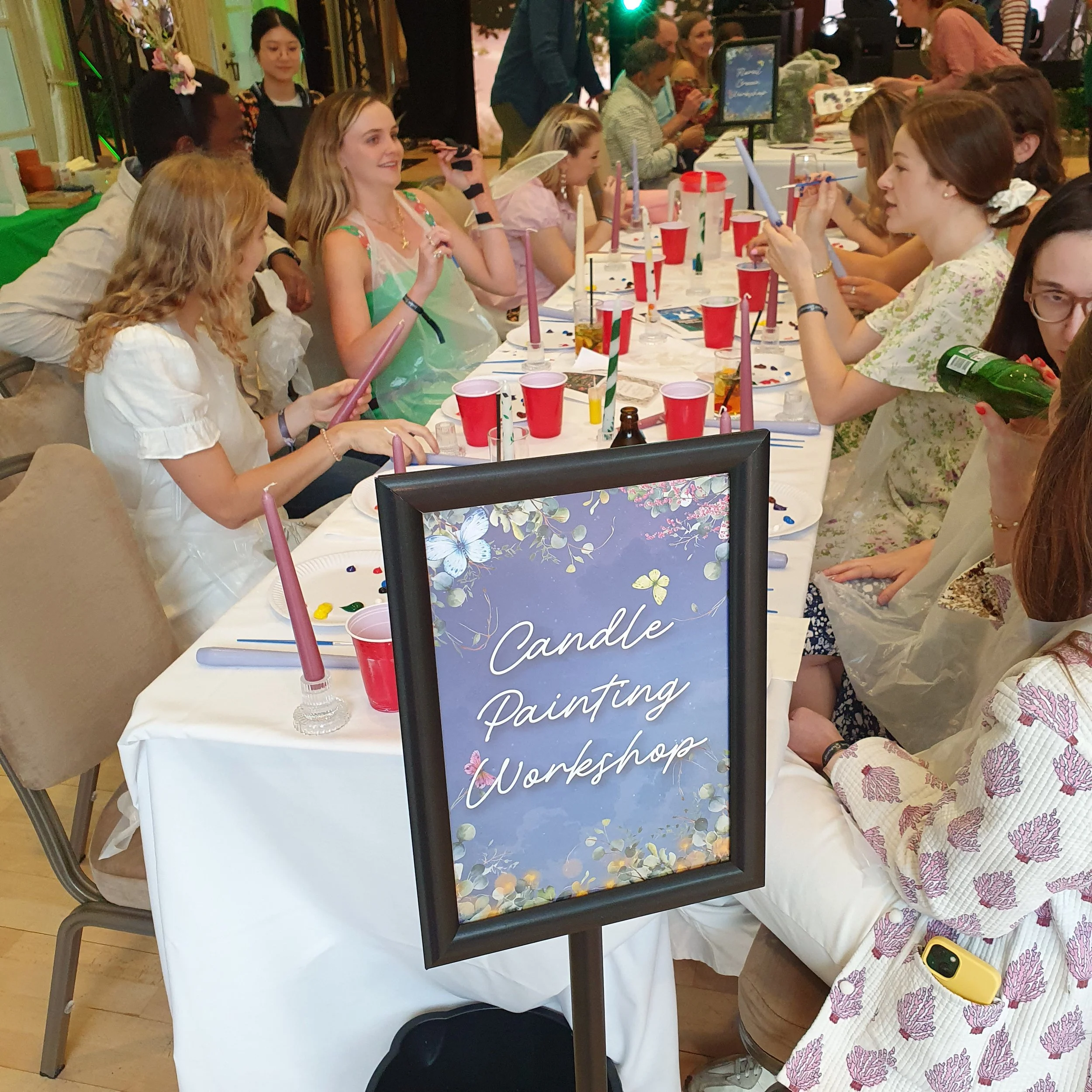 Group of women and children participating in a candle painting workshop, seated around a long table with brushes, candles, and paint. A sign in the foreground reads 'Candle Painting Workshop'.