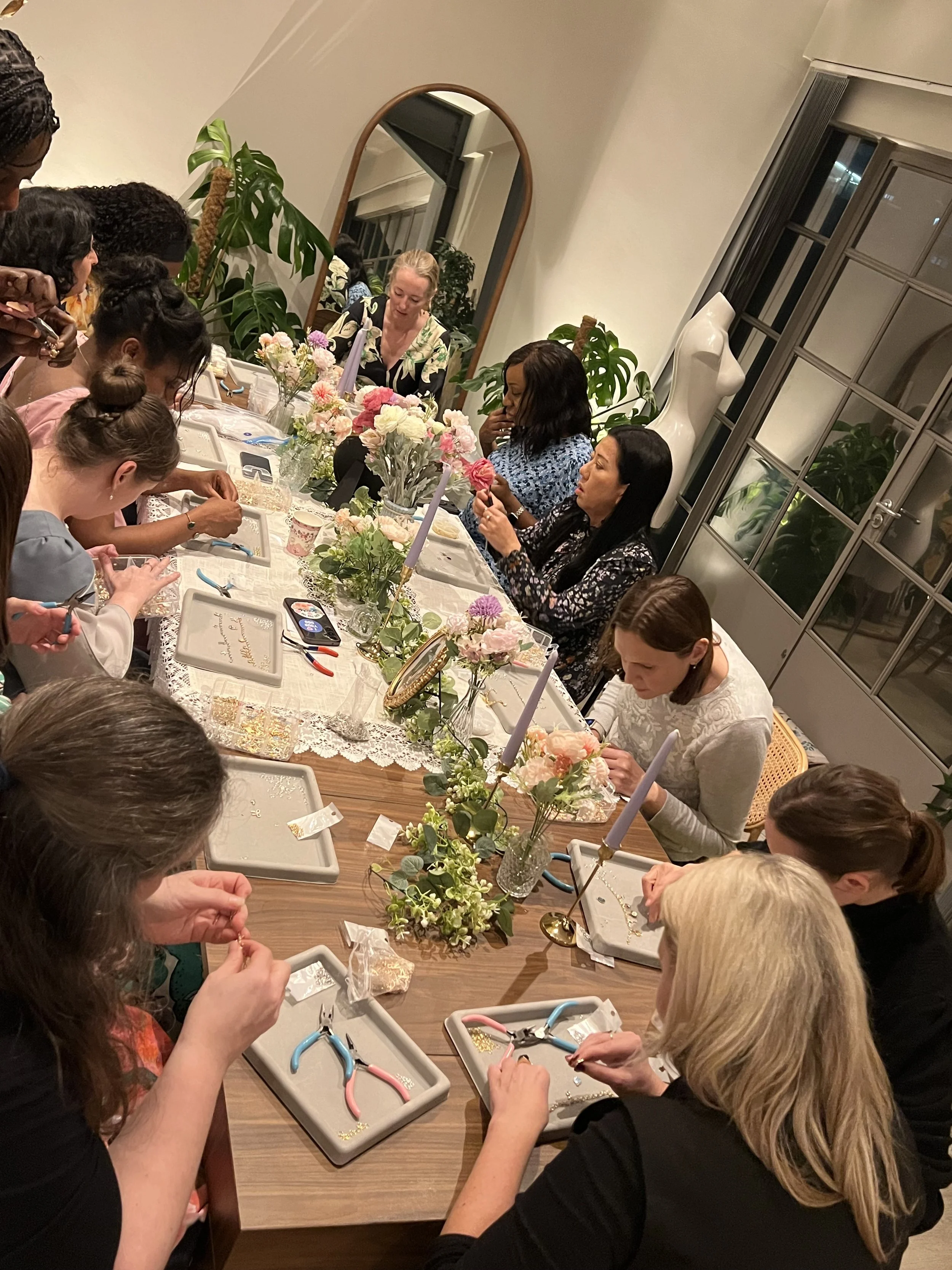 Women sitting around a dining table engaged in jewelry making or crafting activity, with floral centerpieces and candles on the table, during a gathering or workshop in a well-lit room.
