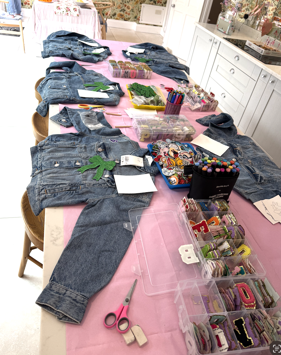 A table with a pink tablecloth displaying denim jackets, patches, craft supplies, fabric markers, scissors, and embroidered patches, likely prepared for customizing or sewing projects.