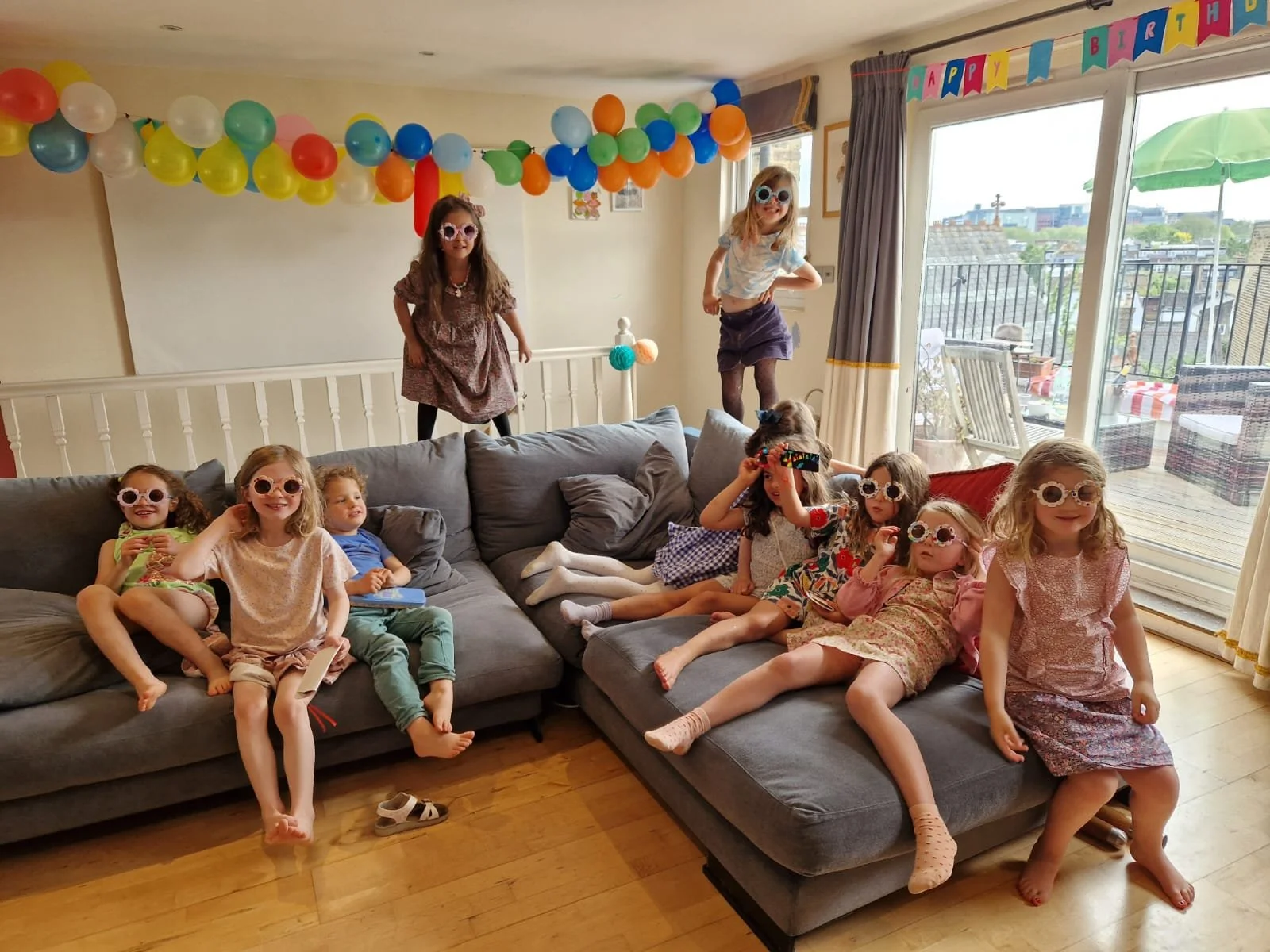 Children celebrating a birthday party in a living room, wearing colorful glasses, with balloons and 'Happy Birthday' banner.