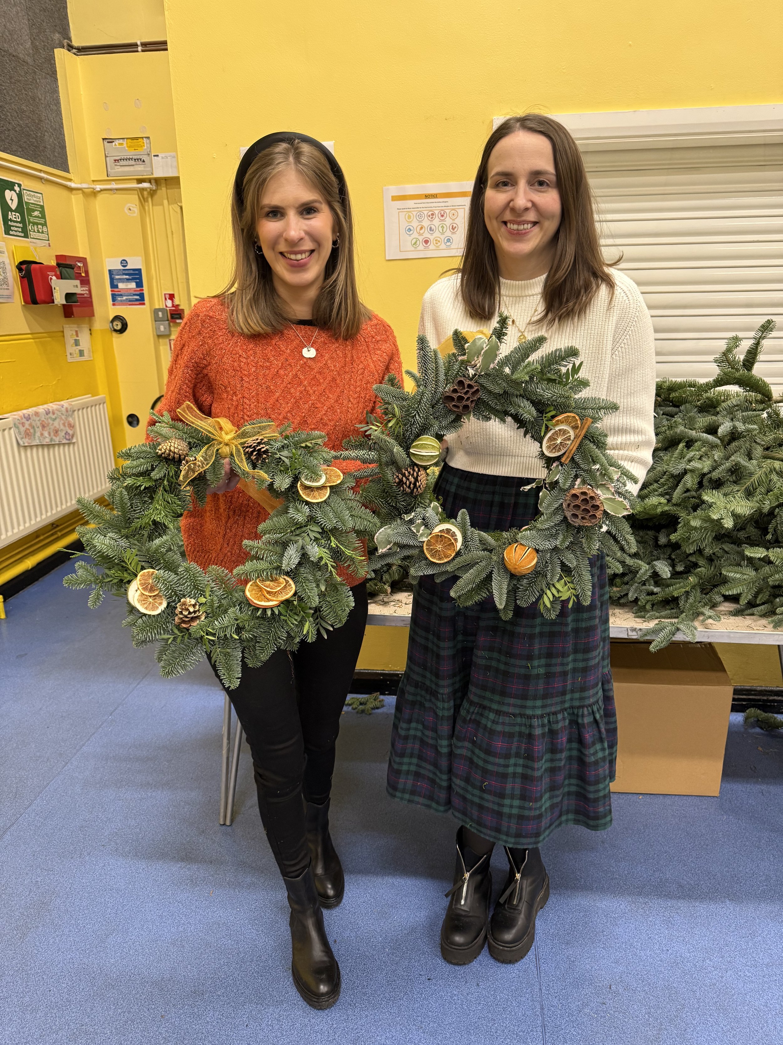 Two women standing indoors smiling, each holding a decorated Christmas wreath made of pine branches with dried orange slices, pine cones, and other natural decorations. They are in front of a table with more pine branches and a yellow wall in the bac