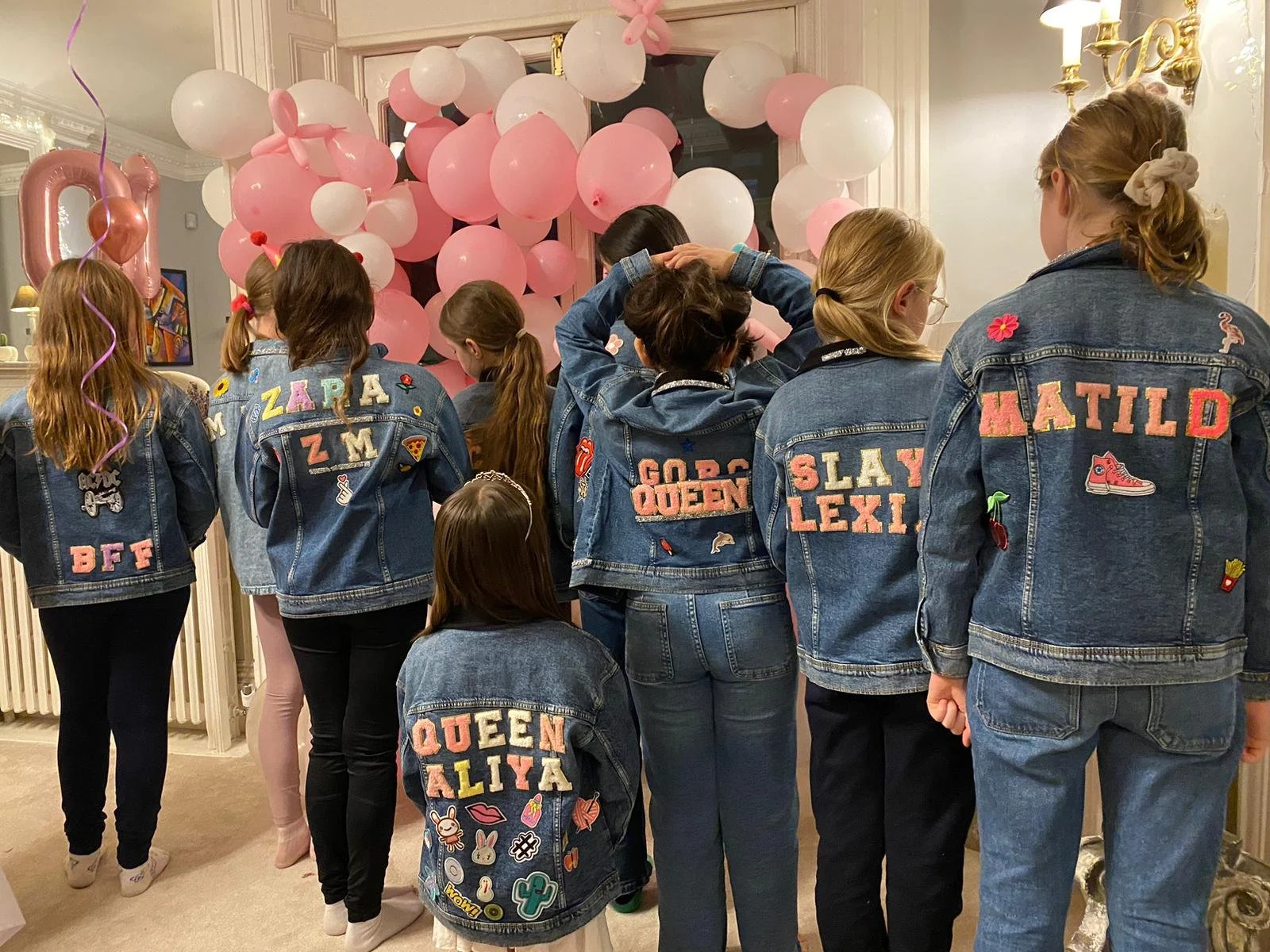 Group of girls wearing denim jackets with custom patches and embroidery, gathered in front of pink and white balloons during a celebration or party.