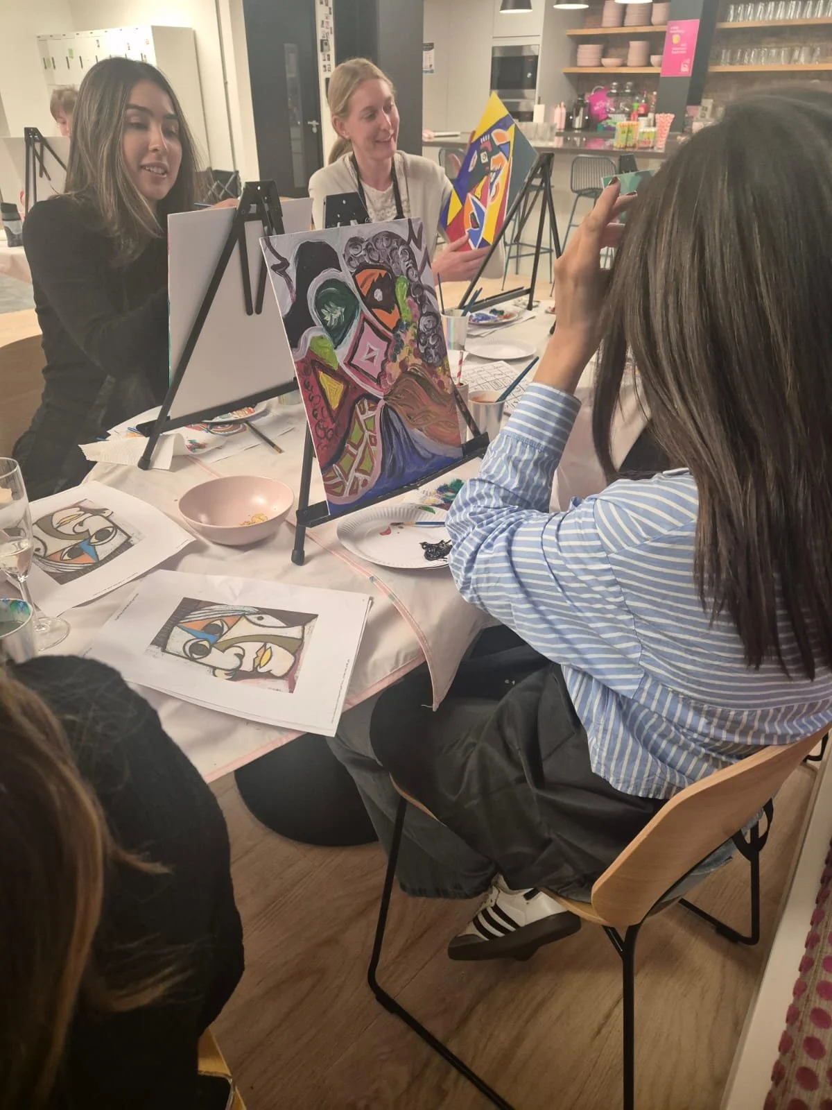 Group of women participating in a painting class, with colorful abstract artworks on display, in a cozy kitchen or cafe setting.