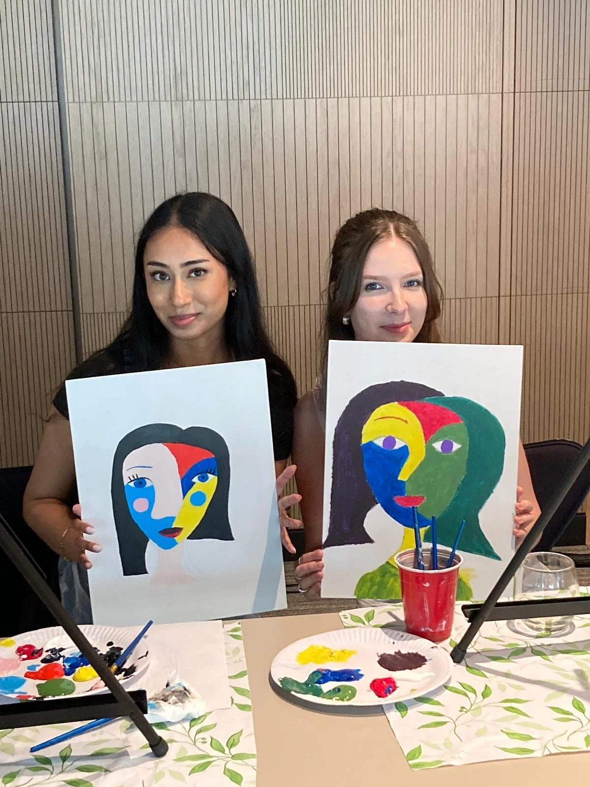 Two women holding abstract colorful portrait paintings of female faces, seated at a table with paint supplies.